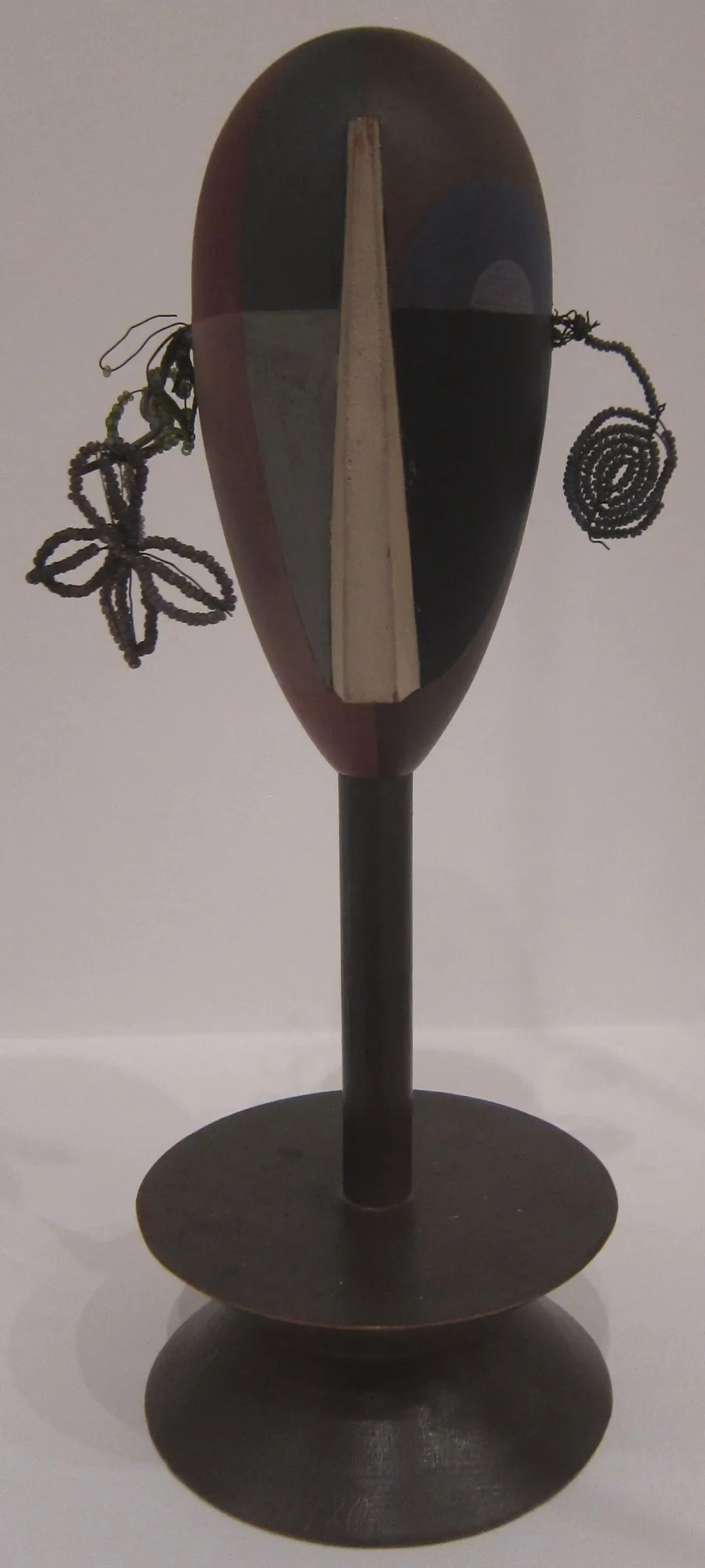 A smooth wooden head shaped like an egg, standing on a turned foot. It is painted in dark bands with a pale stripe down the face, and a loop of bent wire hangs at its side.