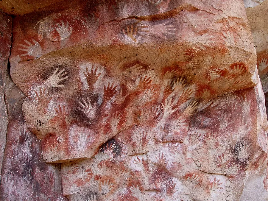 Dozens of hands stencilled onto a rock wall in ochre and white, overlapping each other, all of them left hands, made by many people over a very long time.