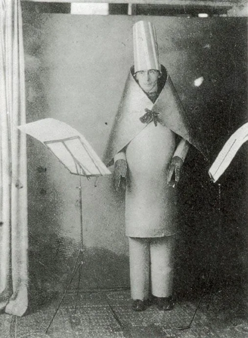 A man stands reading from a music stand, dressed in stiff cardboard tubes for legs, a huge collar and a tall pointed hat. He looks solemn and completely ridiculous at once.